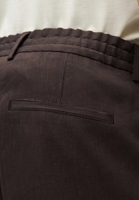 Gros plan sur un pantalon marron foncé avec une taille élastique et une seule poche passepoilée à l'arrière, porté avec un haut blanc.