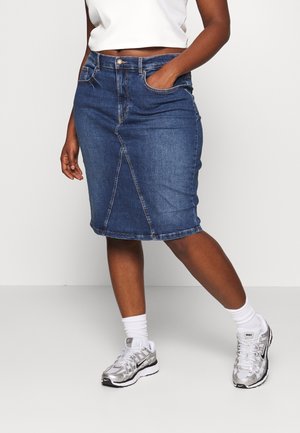 Personne portant une jupe en denim bleu longueur genoux, un top court blanc, des chaussettes blanches et des baskets Nike argentées, debout avec une main dans la poche de la jupe.