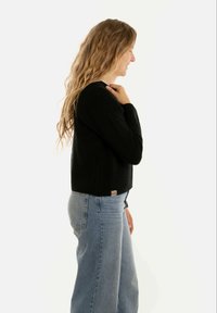 Zwart gebreid cropped trui met lange mouwen; geribbelde textuur en een klein logo-label op de rechterachterkant. Gecombineerd met lichtblauwe jeans.
