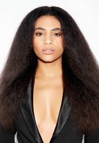 Femme avec des cheveux bouclés naturels volumineux portant un blazer noir en satin à revers en V profond, regardant avec confiance vers l'avant sur un fond blanc.