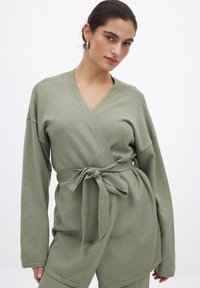 Top avvolgente verde oliva realizzato in tessuto morbido e textured, con scollatura a V profonda e maniche lunghe, legato in vita con una cintura in tessuto.