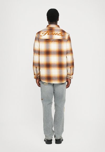 Geltonų apelsinų ir grietinėlės atspalvių plaid ilgomis rankovėmis marškiniai, su siuvinėta logotipu ant nugaros. Suderinta su šviesiai mėlynomis dėvėtomis džinsais.