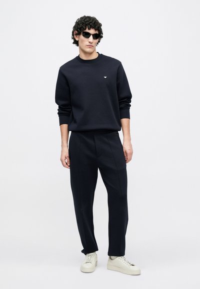 Tmavomodrá mikina s rebrovanými manžetami, skombinovaná s rovnakými plisovanými nohavicami. Obuv tvoria svetlé tenisky. Minimalistický detail loga na hrudi.