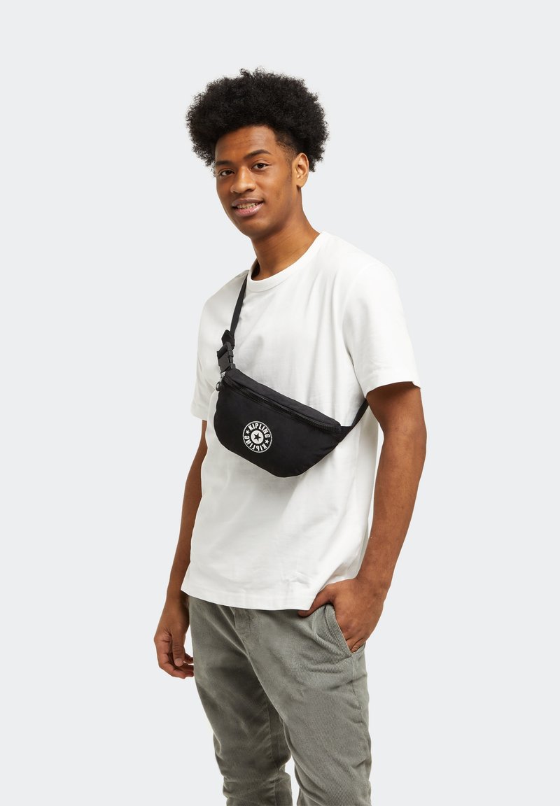 Zwarte stoffen crossbody tas met een gebogen vorm, voorzien van een ritssluiting en een rond logo in het wit. Draag het over een wit shirt.