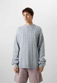 Hellblaues, kabelgestricktes Sweatshirt mit rundem Halsausschnitt und gerippten Bündchen, kombiniert mit hellrosa Shorts. Glatte Textur, lässige Passform.