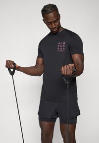 T-shirt de sport noire avec logo rose, assortie à un short. Le mannequin tient des bandes de résistance dans chaque main, mettant en valeur sa musculature.