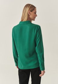 Green long-sleeve shirt made from smooth fabric, featuring a pointed collar and button details on the cuffs, shown from the back.