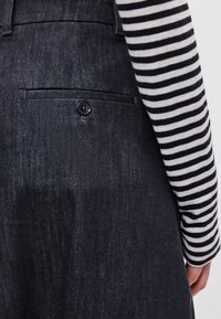 Σκοτεινές τζιν παντελόνες με πίσω τσέπη και μαύρη λεπτομέρεια κουμπιού. Ριγέ μπλούζα μακρύ μανίκι ορατή στα δεξιά. Υφασμάτινη υφή.