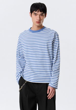 Jeune homme aux cheveux noirs portant une chemise à manches longues rayée bleue et blanche et un pantalon noir avec un accessoire chaîne.