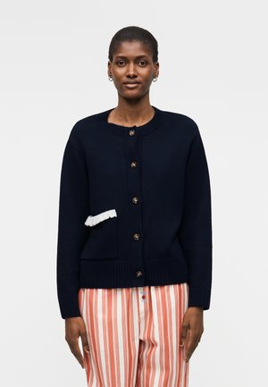 Nő tengerészkék gombos kardigánt visel fehér fodros díszítéssel, valamint narancs-fehér függőlegesen csíkos nadrágot.