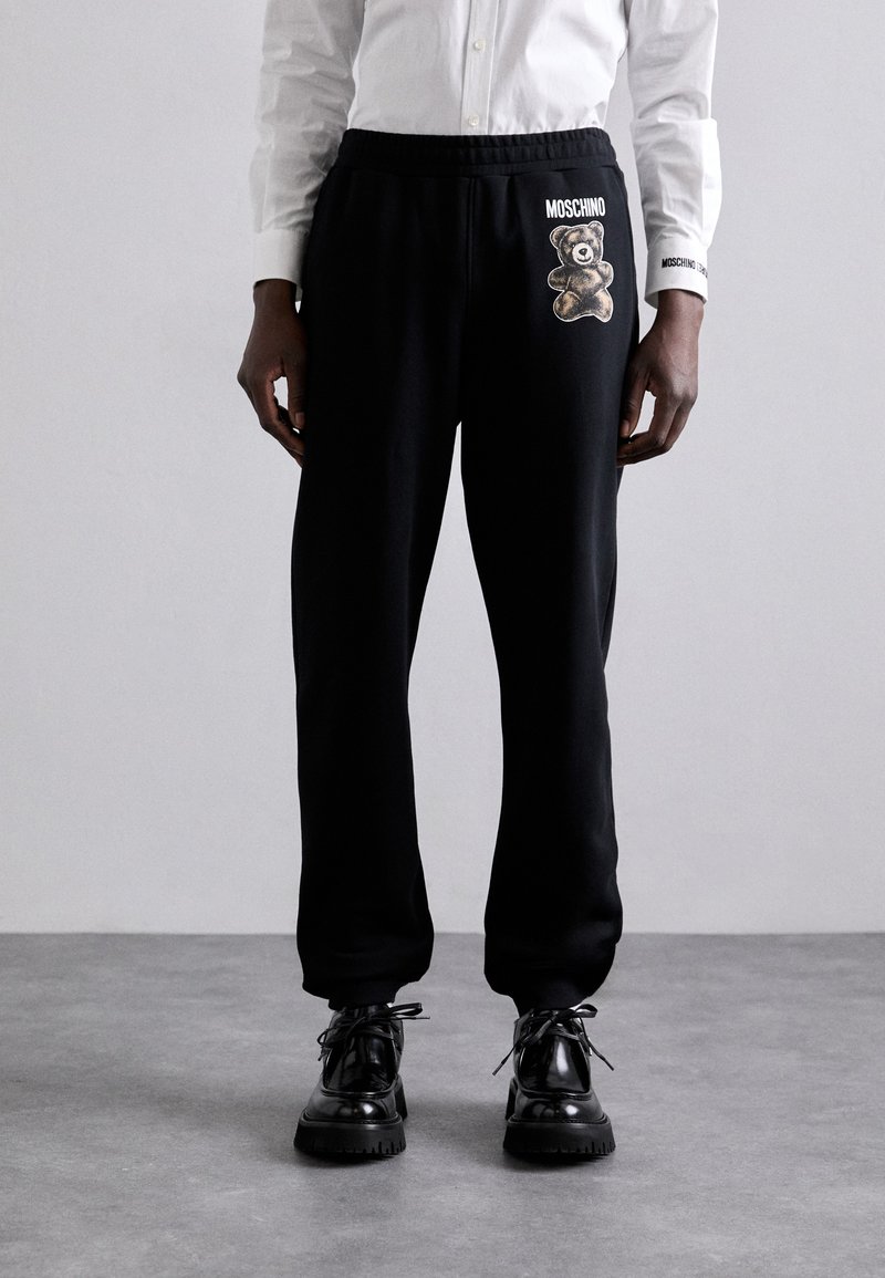 Pantalonii de trening negri au un tiv elastic, un design grafic cu un ursuleț de pluș și accente de logo. Sunt asortați cu pantofi negri lucioși pentru un contrast.