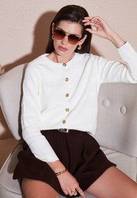 Weißer strukturierter Cardigan mit goldenen Knöpfen, kombiniert mit einem braunen Rock. Das Modell trägt braun getönte Sonnenbrillen und goldenen Schmuck und sitzt auf einem hellen Stuhl.
