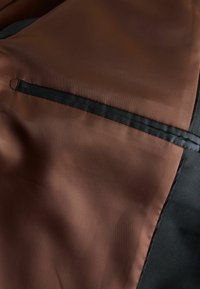 Brauner Stoff mit glatter Textur, ausgestattet mit einer schwarzen Naht und einem kleinen Schlaufen-Detail. Der Hintergrund ist aus schwarzem, geripptem Material.