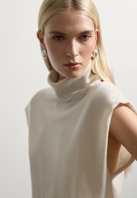Cremfarbener ärmelloser Rollkragenpullover aus weichem Material mit lockerer Passform und seitlichen offenen Nähten, ausgestattet mit angenehmer Textur und minimalistischem Design.