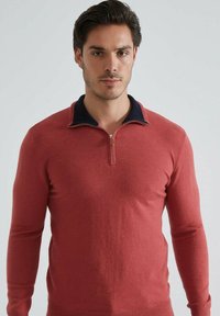 Pull en tricot rouge à manches longues, avec un col zippé et des accents de col bleu marine. Texture lisse, design ajusté et minimalisme dans les détails.