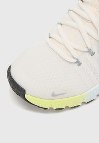 Lichte sneaker met een ademende crèmekleurige bovenkant, grijze veters, een zwarte neus, en levendige gele en blauwe details op de buitenzool.