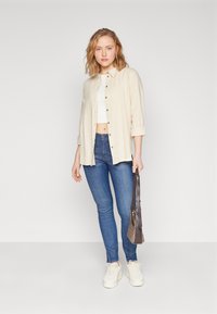 Beige button-up shirt over a white crop top, paired with blue jeans and white sneakers. The model holds a small, patterned bag.