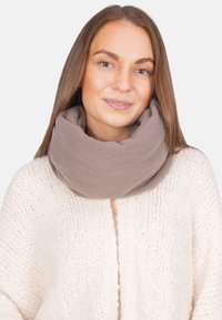 Jeune femme aux cheveux bruns raides portant une écharpe en tricot taupe et un cardigan texturé blanc cassé, souriant doucement.