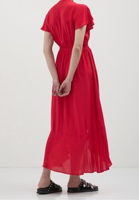 Donna in un vestito rosso alla caviglia con maniche a sbuffo in piedi, con le mani incrociate dietro la schiena, indossa sandali neri con borchie.