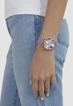 Blumenuhr mit goldfarbenem Gehäuse, pastellviolettem Silikonarmband und lichtreflektierendem silberfarbenem Zifferblatt.