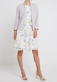 Veste grise claire portée sur une robe fleurie mi-longue aux fleurs jaunes et roses. La robe présente une texture fluide et une ceinture assortie.