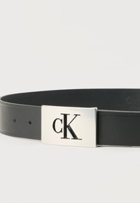 Μαύρη δερμάτινη ζώνη με ορθογώνια ασημί αγκράφα που διαθέτει κομμένα αρχικά "cK" του λογότυπου.