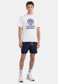 White cotton T-shirt with blue graphic logo, paired with navy shorts. White sneakers and socks with brand name visible.