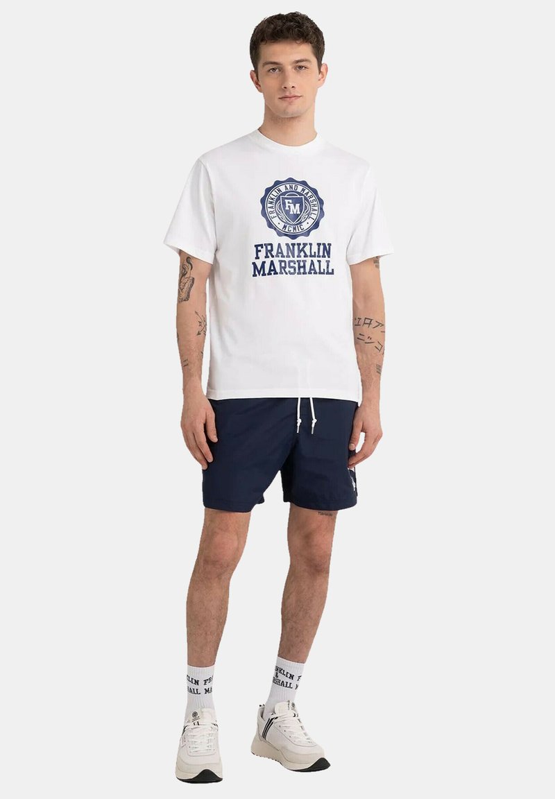 White cotton T-shirt with blue graphic logo, paired with navy shorts. White sneakers and socks with brand name visible.