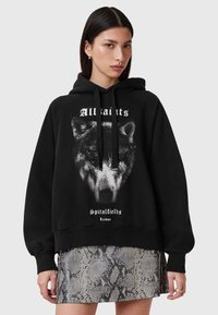 Sudadera negra de corte oversize que presenta un gráfico de un lobo en el frontal, cordones ajustables y texto de "All Saints", combinada con una falda plateada con patrón de piel de serpiente.