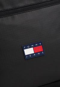 Musta, teksturoidusta kankaasta valmistettu esine, jossa näkyy vetoketju sekä suorakulmainen sininen merkki, jossa lukee "TOMMY JEANS" ja punaisia, valkoisia ja sinisiä osioita.