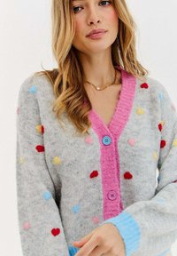 Szary dzianinowy kardigan ozdobiony kolorowymi wzorami serc, z różowym prążkowanym kołnierzem, niebieskimi guzikami i niebieskimi mankietami.