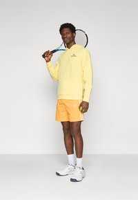 Gul hættetrøje, orange atletiske shorts med striber, hvide sneakers, holder en tennisracket. Glat stof, afslappet pasform.