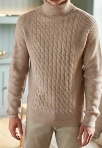 Suéter beige de cuello alto tejido con diseño texturizado de trenzas en la parte delantera, mangas y borde acanalados. Material suave similar a la lana.