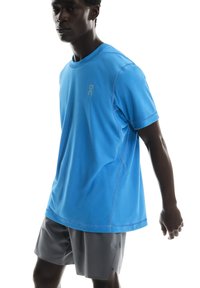 T-shirt de sport bleu à manches courtes et col rond, avec un logo discret. Associé à un short gris, mettant en avant un ajustement confortable.