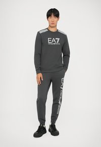 Felpa grigia con logo EA7 e accenti a strisce sulle spalle, abbinata a pantaloni della tuta coordinati con testo EA7, indossati con scarpe da ginnastica nere.