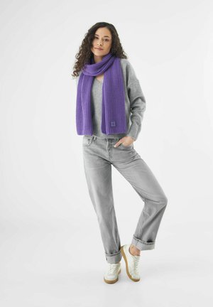 Femme aux cheveux bouclés portant un pull gris, une écharpe violette, un jean gris et des baskets blanches, se tenant avec une main dans la poche contre un fond blanc.