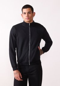 Čierna mikina na zips s vysokým golierom, vyrobená z mäkkej tkaniny. Má rebrované manžety a spodný lem, ako aj bočné vrecká. Hladká štruktúra, minimalistický dizajn.