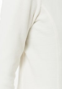 Primer plano de una prenda de tela acolchada blanca que muestra la manga y el panel lateral con detalles de costura horizontal.