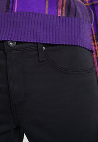 Top de suéter de canalé morado sobre jeans ajustados negros, con cierre de botón y bolsillos delanteros. La tela parece suave con un sutil estiramiento.