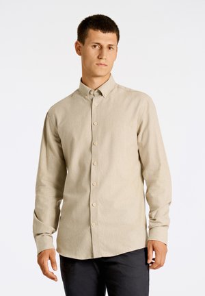 Chemise à boutons beige clair en tissu texturé, avec un col et des manches longues. Détails de couture subtils visibles.