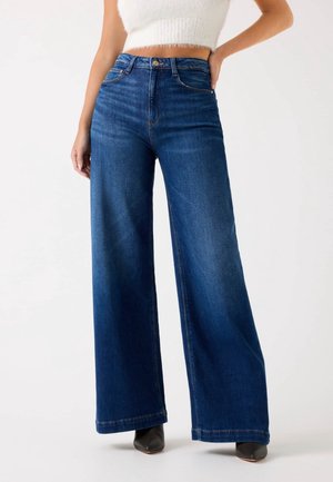 Kobieta nosząca ciemnoniebieskie, szerokie w nogawkach jeansy z wysokim stanem, zestawione z czarnymi botkami z ostrym noskiem i białym, krótkim, bezrękawnym topem.