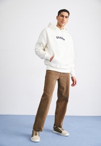 Sudadera crema con el logo de "Dickies", pantalones de pana marrón y zapatillas beige-verdes, de pie sobre un suelo azul contra una pared clara.