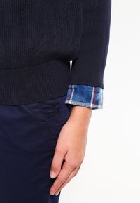 Granatowy sweter dzianinowy z teksturowymi wzorami, z widocznymi podwiniętymi mankietami niebieskiej kraciastej koszuli. Ciemnogranatowe spodnie z boczną kieszenią.