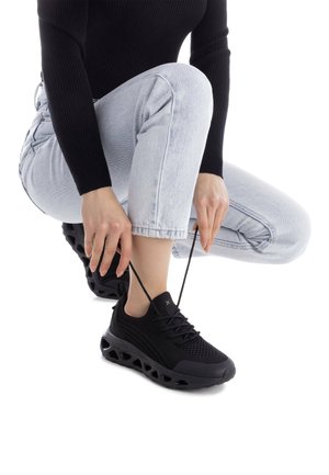 Zapatillas deportivas negras con parte superior de malla, acentos texturizados y una suela de espuma esculpida. Se están ajustando los cordones, combinadas con unos pantalones vaqueros gris claro.