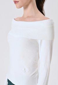 Frau in einem weißen, schulterfreien, gerippten Strickpullover mit langen Ärmeln, sichtbaren Schlüsselbeinen und dunkelgrünen Hosen vor einem einfarbigen Hintergrund.