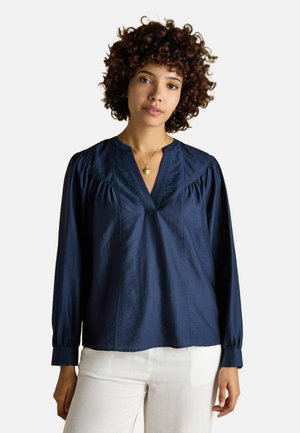 Woman with curly hair wearing a navy blue long-sleeve blouse with a v-neck and white pants against a plain background.