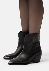 Botas de vaquero negras de cuero hasta el tobillo con punta afilada y tacón medio cuadrado, con costuras curvadas y lengüetas para facilitar el uso.