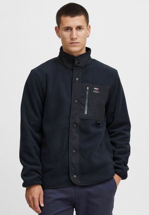 Chaqueta de forro polar azul oscuro con un alto cuello, bolsillo en el pecho con cremallera, cierre a presión y textura suave. Presenta acentos en negro contrastante.