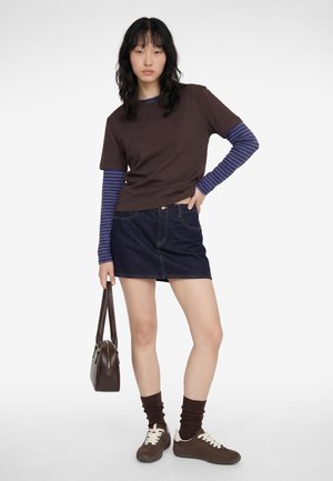 Ung kvinde iført brun t-shirt over blåstribet langærmet trøje, denimskørt, brune sneakers, matchende sokker, og holder en brun håndtaske.