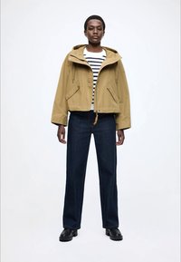 Χακί cropped μπουφάν με κουκούλα και κορδόνι, συνδυασμένο με σκούρο μπλε τζιν κανονικής γραμμής. Κάτω από αυτό, λευκό πουκάμισο με μπλε ρίγες. Μαύρα παπούτσια.